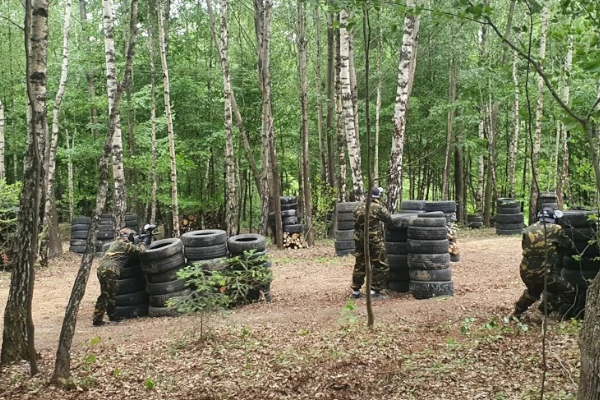 Trzej gracze w maskach i strojach moro biorą udział w grze w paintball na leśnym polu ze stosami opon jako osłonami.