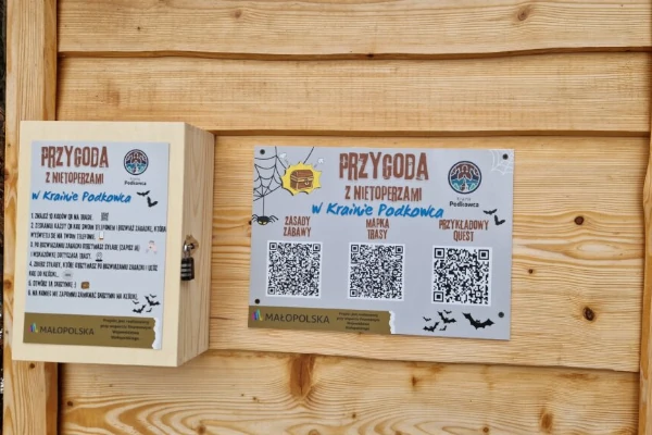 Informacyjna tablica drewniana dotycząca gry terenowej „Przygoda z nietoperzami w Krainie Podkowca”, zawierająca instrukcję i kody QR.